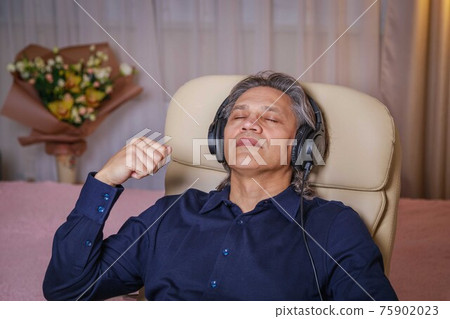 50-year-old man listens to music on headphones at home, sitting in a chair. 50-year-old man listens to music on headphones at home, sitting in a chair. 75902023