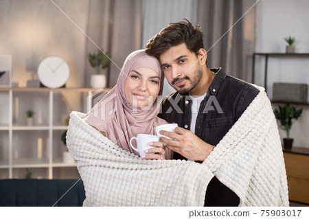 Family portrait indoors. Close up of happy lovely Muslim woman in hijab and handsome bearded Arabian man, standing at home living room, covered with warm plaid and drinking hot coffee together 75903017