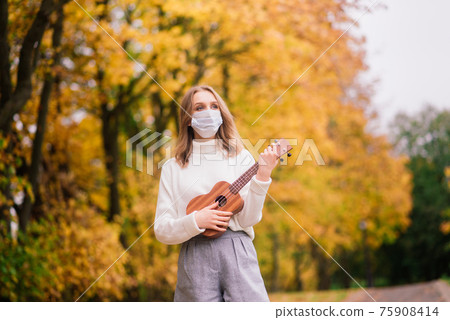 Portrait young woman in protective mask playing ukulele guitar in autumn park, healthy travel 75908414
