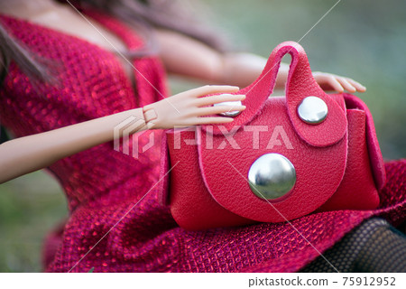 Closeup of mini red leather hand bag in hands of mannequin doll sitting in the grass in a public garden 75912952