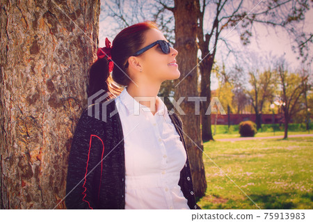 Young girl with sunglasses leaning against a tree in the park Young girl with sunglasses leaning against a tree in the park 75913983