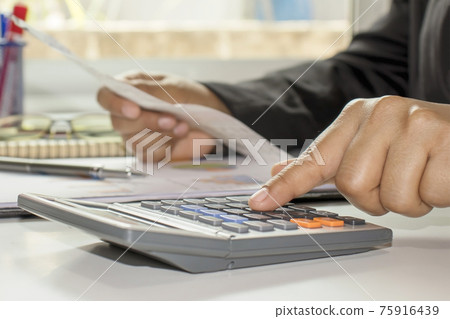 The businessman's hand is pressing a calculator, doing finance work and calculating on the table about expenses at home office. The businessman's hand is pressing a calculator, doing finance work and calculating on the table about expenses at home office. 75916439