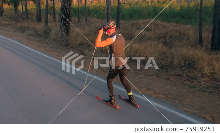 Training an athlete on the roller skaters. Biathlon ride on the roller skis with ski poles, in the helmet. Autumn workout. Roller sport. 75919251