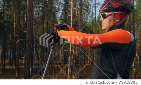 Training an athlete on the roller skaters. Biathlon ride on the roller skis with ski poles, in the helmet. Autumn workout. Roller sport. Athlete is getting ready to start. 75919293