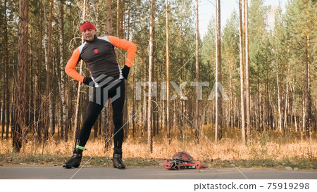 Training an athlete on the roller skaters. Biathlon ride on the roller skis with ski poles, in the helmet. Autumn workout. Roller sport. The athlete does the workout. 75919298