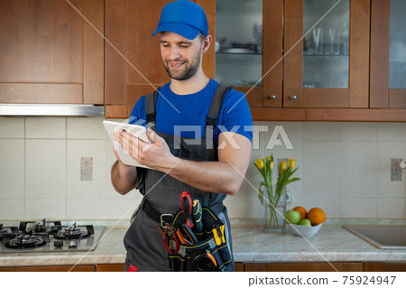 Plumber wearing a tool belt with various tools using tablet during work in kitchen Plumber wearing a tool belt with various tools using tablet during work in kitchen 75924947