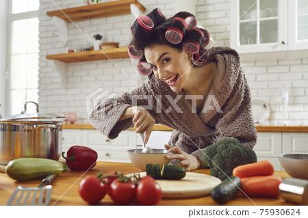 Funny woman housewife in curlers and bathrobe standing cooking fresh healthy dish Funny woman housewife in curlers and bathrobe standing cooking fresh healthy dish 75930624