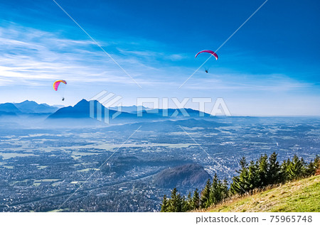 Paragliding over Salzburg in Austria, Europe. Two paragliders, seen from 1287 meters high Gaisberg mountain, with view of capital city of Salzburg, the fortress Hohensalzburg and vicinity. Photo. Paragliding over Salzburg in Austria, Europe. Two paragliders, seen from 1287 meters high Gaisberg mountain, with view of capital city of Salzburg, the fortress Hohensalzburg and vicinity. Photo. 75965748