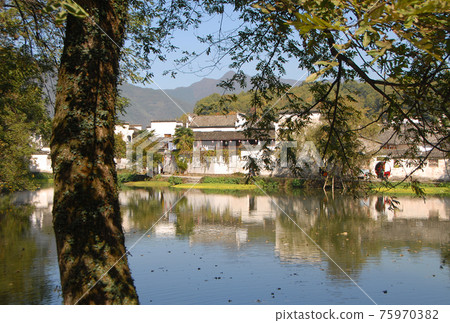 Hongcun Ancient Town in Anhui Province, China. Landscape view of the old buildings of Hongcun with their reflection in the water of Nanhu Lake and foreground tree. The ancient town of Hongcun in China Hongcun Ancient Town in Anhui Province, China. Landscape view of the old buildings of Hongcun with their reflection in the water of Nanhu Lake and foreground tree. The ancient town of Hongcun in China 75970382