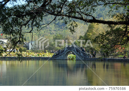 Hongcun Ancient Town in Anhui Province, China. Landscape view of the stone bridge crossing Nanhu Lake in Hongcun. Framed by foreground branches. The ancient town of Hongcun in China Hongcun Ancient Town in Anhui Province, China. Landscape view of the stone bridge crossing Nanhu Lake in Hongcun. Framed by foreground branches. The ancient town of Hongcun in China 75970386