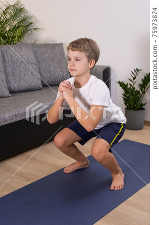 Blond boy goes in for sports on yoga mat. Child physical activity squat exercise at home. 75973874