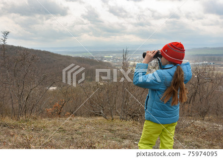 A girl examines a mountain landscape through binoculars, view from the back A girl examines a mountain landscape through binoculars, view from the back 75974095
