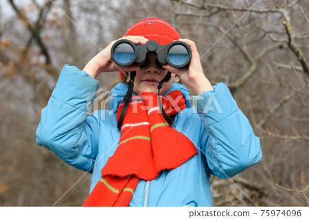 A girl looks through binoculars on a background of a winter forest, front view, close-up A girl looks through binoculars on a background of a winter forest, front view, close-up 75974096
