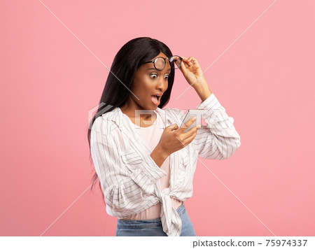 Shocked black lady looking at smartphone in disbelief, lifting her eyeglasses on pink studio background 75974337