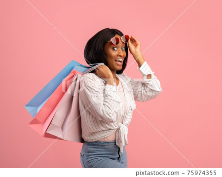 Pretty black woman with shopping bags lifting sunglasses, feeling excited on pink studio background Pretty black woman with shopping bags lifting sunglasses, feeling excited on pink studio background 75974355