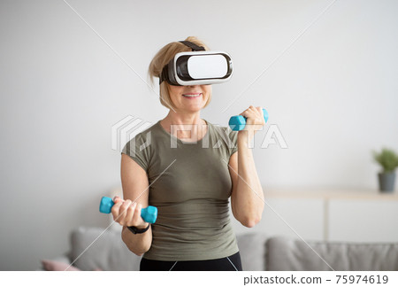 Sporty mature woman in VR headset working out her biceps with dumbbells during domestic workout Sporty mature woman in VR headset working out her biceps with dumbbells during domestic workout 75974619