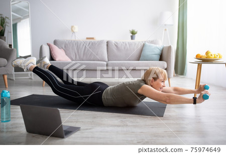Strong mature woman making exercises with dumbbells on yoga mat, working out to video tutorial at home 75974649