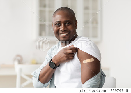 Covid-19 vaccination program. Positive black man holding up shirt sleeve showing plaster after coronavirus vaccine shot Covid-19 vaccination program. Positive black man holding up shirt sleeve showing plaster after coronavirus vaccine shot 75974746