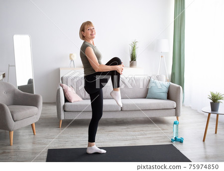 Happy senior woman doing flexibility exercises, stretching her leg on yoga mat at home, full length 75974850