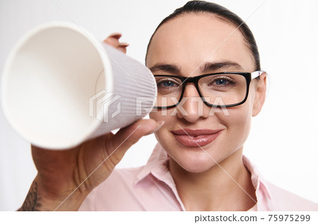 Adorable brunette woman in eyeglasses looks smiling at camera through takeaway paper cup. Closeup, white background, copy space 75975299