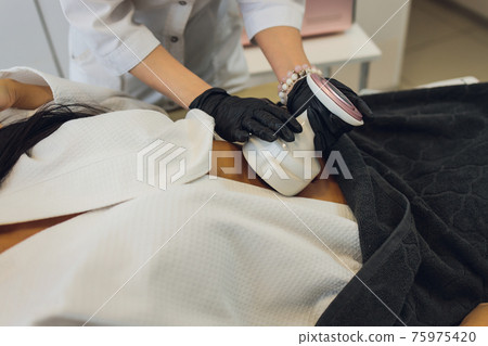 Woman in special white suit getting anti cellulite massage in a spa salon. LPG, and body contouring treatment in clinic. Woman in special white suit getting anti cellulite massage in a spa salon. LPG, and body contouring treatment in clinic. 75975420
