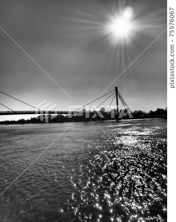 Bridge over the Sava river in Sremska Mitrovica, Serbia. Black and white monochrome photography in retro and vintage style. Reflections of rays on the water. Full-flowing river. Sun in the sky. Bridge over the Sava river in Sremska Mitrovica, Serbia. Black and white monochrome photography in retro and vintage style. Reflections of rays on the water. Full-flowing river. Sun in the sky. 75976067