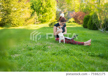 Caucasian woman is typing on a laptop while sitting on a green lawn outdoors. Female freelancer works remotely in the park. 75982175