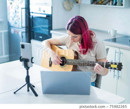 A woman sits in the kitchen during a remote acoustic guitar lesson. A girl learns to play the guitar and watches educational videos on a laptop 75982265