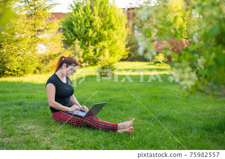 A red-haired woman sits in green grass in a park and is shopping online. The girl maintains a social distance and works outdoors on a laptop. A student is studying remotely on a computer. A red-haired woman sits in green grass in a park and is shopping online. The girl maintains a social distance and works outdoors on a laptop. A student is studying remotely on a computer. 75982557