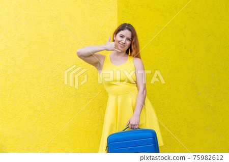 A beautiful red-haired woman in a dress holds a blue suitcase and gestures a phone call on a yellow background. 75982612