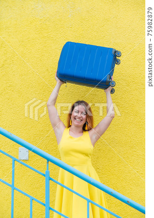 Happy woman in a yellow dress holds a blue suitcase over her head against the background of a yellow wall. Caucasian girl smiles with teeth and happily waits for a trip. Happy woman in a yellow dress holds a blue suitcase over her head against the background of a yellow wall. Caucasian girl smiles with teeth and happily waits for a trip. 75982879