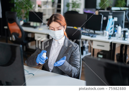 Female manager is sitting in a chair at the workplace. A business woman in a mask is spraying gloves with an antiseptic. Concept of office work during the coronavirus epidemic. Personal hygiene. Female manager is sitting in a chair at the workplace. A business woman in a mask is spraying gloves with an antiseptic. Concept of office work during the coronavirus epidemic. Personal hygiene. 75982880
