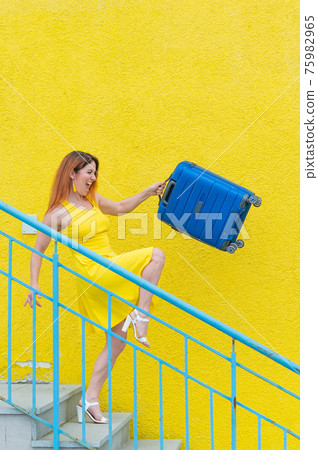 A happy red-haired woman in a yellow dress is walking down the stairs and joyfully swing a blue suitcase. The girl is preparing for the journey. Summer vacation concept. 75982965