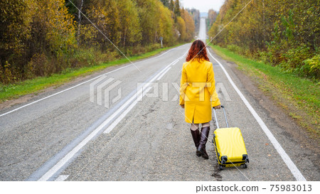 A red-haired woman in a yellow coat walks with a yellow suitcase along the highway in autumn. A red-haired woman in a yellow coat walks with a yellow suitcase along the highway in autumn. 75983013