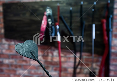 A set of varied leather lashes for sexual pleasures hang on a wooden hanger on a brick wall. Sex equipment in red and black for role-playing games. BDSM room. A set of varied leather lashes for sexual pleasures hang on a wooden hanger on a brick wall. Sex equipment in red and black for role-playing games. BDSM room. 75983014