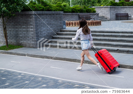 Happy beautiful caucasian woman is running with a big red suitcase outdoors. Excited girl is late for the plane. 75983644