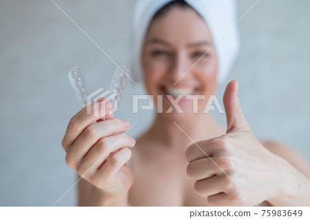 Orthodontic silicone transparent teeth aligner in female hands. A blurred woman with a towel on her head holds a removable retainer and shows a thumb up. Bracket for teeth whitening. Dental care. 75983649