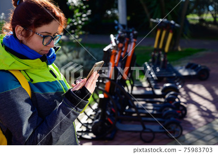 The girl in the jacket uses the mobile application to pay and unlock the rental electric scooter. Close-up female hand with a phone. Woman on eco-friendly transport to move around the city. 75983703