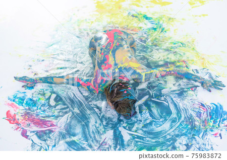 Top view of a naked woman in a spray of paint. A girl without clothes lies on a white floor in colored spots. Original body painting. 75983872
