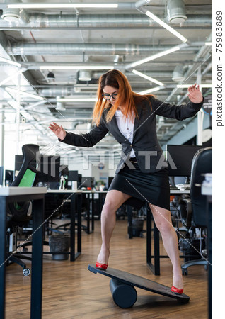 A woman in a suit is engaged on a rocker-skateboard during a break at work. A female office employee in high heels stands on a balance board. A woman in a suit is engaged on a rocker-skateboard during a break at work. A female office employee in high heels stands on a balance board. 75983889