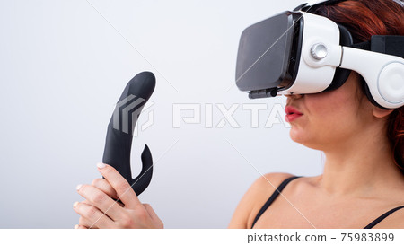 Caucasian red-haired woman wearing virtual reality glasses holding black dildo and looking lustfully. Caucasian red-haired woman wearing virtual reality glasses holding black dildo and looking lustfully. 75983899