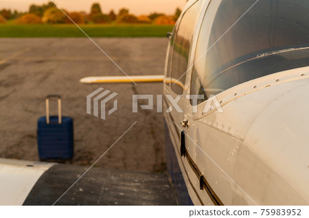 A blue suitcase and a parked small private jet. Quadruple plane with a propeller for an air taxi in the sunset. Self travel concept. plane for VIP persons. A blue suitcase and a parked small private jet. Quadruple plane with a propeller for an air taxi in the sunset. Self travel concept. plane for VIP persons. 75983952