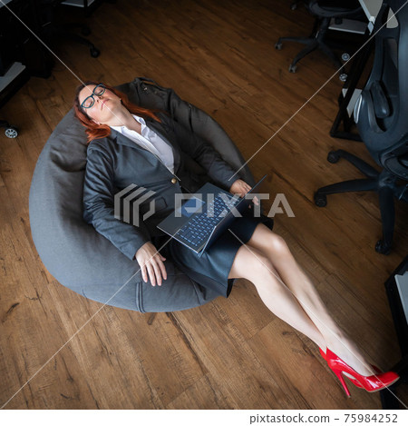 Modern advanced office. A woman in a business suit asleep in a beanbag while working on a laptop. Female manager is resting in a soft chair. Caring for the comfort of employees. 75984252