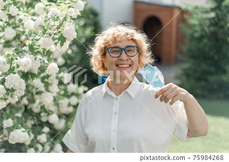 Happy old woman takes off the mask and sniffs a blossoming apple tree. A female pensioner enjoys fresh air on a warm summer day in the garden. Quarantine is over. The end of the coronavirus pandemic. Happy old woman takes off the mask and sniffs a blossoming apple tree. A female pensioner enjoys fresh air on a warm summer day in the garden. Quarantine is over. The end of the coronavirus pandemic. 75984268
