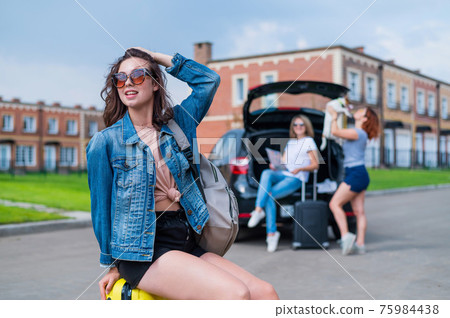 Three girlfriends go on a road trip. Young woman posing with a suitcase. Two girls are sitting on the trunk of a car and watching a map. 75984438