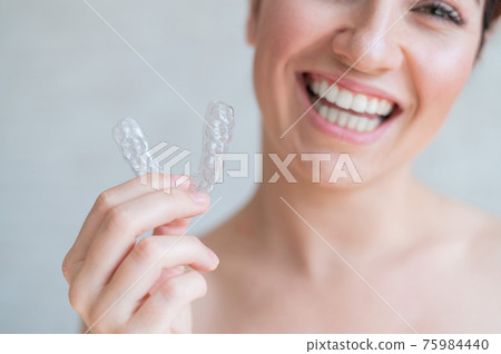 Close-up of orthodontic silicone transparent teeth aligner in female hands. A woman with a perfect charm smile holds a removable night retainer. Bracket for teeth whitening. Cropped photo. Close-up of orthodontic silicone transparent teeth aligner in female hands. A woman with a perfect charm smile holds a removable night retainer. Bracket for teeth whitening. Cropped photo. 75984440