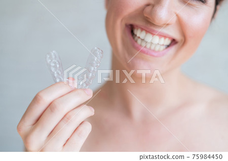 Close-up of orthodontic silicone transparent teeth aligner in female hands. A woman with a perfect charm smile holds a removable night retainer. Bracket for teeth whitening. Cropped photo. 75984450