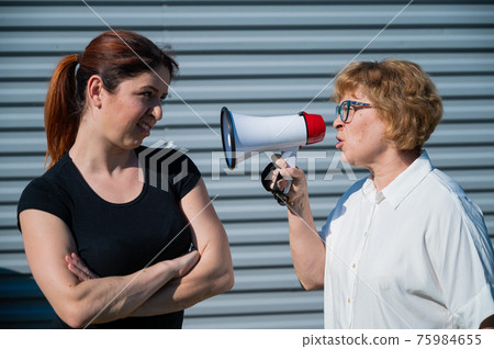 The conflict of generations. An old woman yells into a megaphone at her middle-aged daughter against gray background. Quarrel between an elderly mother and daughter. The conflict of generations. An old woman yells into a megaphone at her middle-aged daughter against gray background. Quarrel between an elderly mother and daughter. 75984655