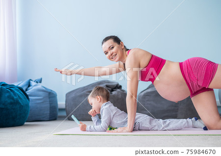 A pregnant woman in sportswear does gymnastics while her son plays in the mobile. Girl on yoga with her child. Fitness mom and baby. 75984670