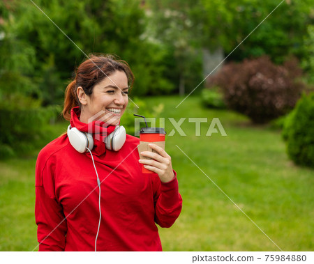 Red-haired woman with headphones and drinks coffee with a straw in the park. Girl enjoys a walk on a sunny summer day and holds a takeaway cappuccino cup. Headphones connected to a smartphone. Red-haired woman with headphones and drinks coffee with a straw in the park. Girl enjoys a walk on a sunny summer day and holds a takeaway cappuccino cup. Headphones connected to a smartphone. 75984880
